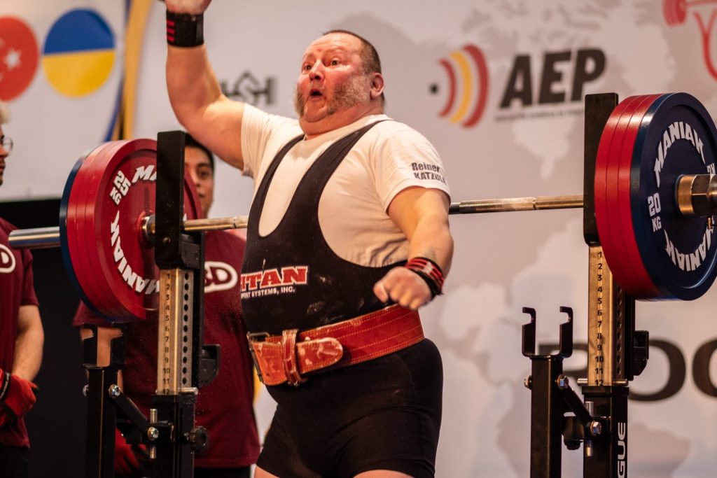 Kraftdreikämpfer im Wettkampfanzug und rotem Gürtel jubelt nach einem erfolgreichen Versuch und streckt den Arm in die Höhe