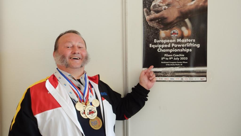 Lachender Kraftsportler mit Goldmedaillen und Sportjacke zeigt auf ein Powerlifting-Meisterschafts-Poster von 2023